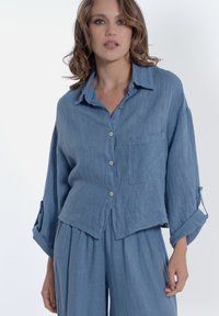 Camicia in lino azzurro chiaro con vestibilità rilassata, chiusura con bottoni e taschino singolo sul petto. Caratterizzata da maniche arrotolate con bottoni. 