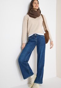 Pull beige à manches longues, jean bleu à jambes larges, écharpe marron et baskets crème. Le mannequin est debout contre un mur blanc, tenant un sac à bandoulière marron.