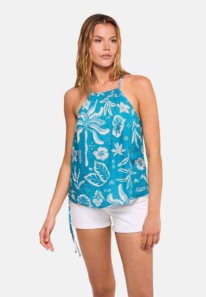 Mujer con top azul de cuello halter con estampado tropical blanco y shorts blancos, de pie frente a un fondo liso y claro.
