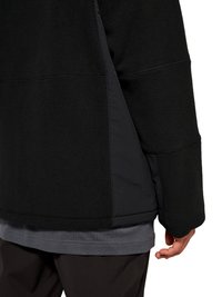 Close-up af en persons ryg iført en sort fleecejakke over en grå trøje og mørke bukser, med højre hånd afslappet ved siden.