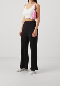 Pantalon large côtelé noir avec un crop top noir ajusté et des baskets blanches. Un petit sac à main rose est porté en bandoulière sur une épaule.
