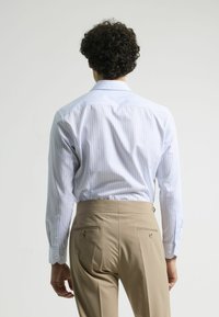 Camisa a rayas azul claro de mangas largas, vista trasera. Combinada con pantalones beiges de corte tailored con bolsillos laterales y una textura suave.