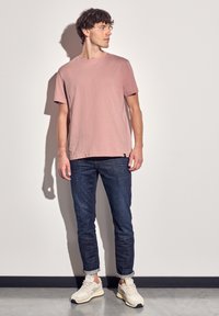 Camiseta de cuello redondo en rosa claro, combinada con jeans oscuros de corte ajustado, y zapatillas beige. Los jeans están ligeramente doblados en los puños.