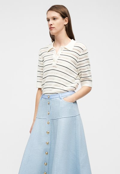 Γυναίκα που φοράει πλεκτό τοπ με κοντά μανίκια σε λευκό και μαύρο ριγέ, που είναι μπουζωμένο μέσα σε φούστα ψηλόμεση ανοιχτό μπλε με κουμπιά μπροστά και τσέπες.