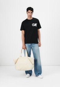 T-shirt de algodão preta com logotipo branco, combinada com jeans de perna larga azul claro. O modelo segura uma bolsa de lona bege com detalhe de logotipo em relevo.