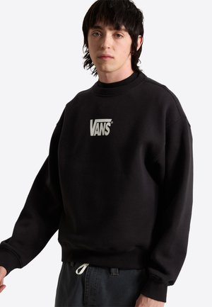 Ung mand med mørkt hår iført en sort Vans sweatshirt og mørke bukser mod en ensfarvet hvid baggrund.