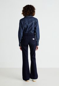 Mörka denimbyxor med utsvängd nederdel, som matchas med en denimskjorta med tryckknappar och en yoke-design, som visar en klassisk westernstil.