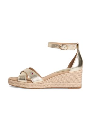 Sandalia de cuña metálica dorada con correa en el tobillo y hebilla, plataforma de yute trenzado y pequeño logo de bandera en la correa frontal.