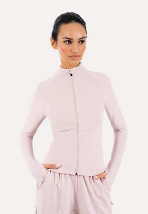 Mujer con cabello oscuro con una chaqueta rosa pálido con cremallera y pantalones a juego, manos en los bolsillos, mirando ligeramente hacia un lado.