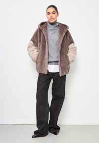 Giacca in pelliccia sintetica color tortora e beige, con zip e cappuccio. Indossata sopra un maglione grigio a coste e un paio di jeans scuri larghi, con scarpe nere a punta.