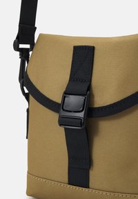 Tan messenger bag featuring a textured fabric, black adjustable strap, and a buckle closure with detailed stitching along the bottom edge.