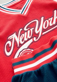 Červený sieťovaný dres s nápisom "New York" v bielej farbe, orámovaný modrou. Obsahuje tmavozelenú časť šortiek so pruhovaným pásom v červenej, bielej a modrej farbe.