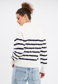 Pull rayé blanc et marine avec un col, des poignets et un ourlet côtelés, porté avec un jean bleu clair. Texture en tricot douce.