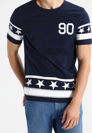 Marineblaue Kurzarmbluse mit weißen Sternmustern an Ärmeln und Saum sowie der weißen Zahl "90" auf der Brust, kombiniert mit blauen Jeans.