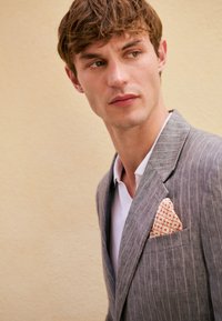 Blazer grigio a righe sottili con rever a incavo, completato da un fazzoletto da taschino arancione fantasia. Camicia bianca sotto, sfondo chiaro.