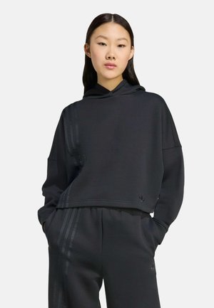 Felpa nera a maniche lunghe, realizzata in tessuto morbido, con cappuccio e tre strisce tonali su un lato. Abbinata a pantaloni joggers scuri coordinati.