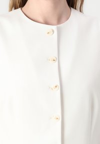Hvit bluse laget av glatt stoff, med rund hals og fem kremfargede knapper foran som utpreget detalj.