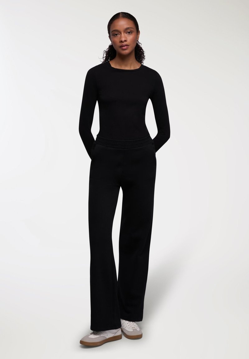 Maglietta nera a maniche lunghe abbinata a pantaloni neri a gamba larga. L'outfit presenta una consistenza liscia e una cintura elastica. Sneakers bianche casual completano il look.