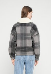 Jachetă gri cu model în carouri și guler din blană de oaie, cu croială lejeră și manșete cu nasturi, combinată cu blugi bleu cu dungi verticale.