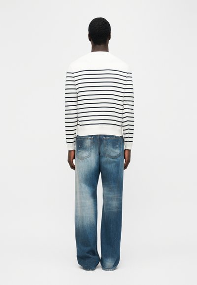 Homme portant une chemise blanche à manches longues avec des rayures horizontales noires et un jean large bleu délavé, debout de dos sur un fond uni.