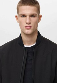 Musta pommitakki, jossa on sileä pinta ja pyöreä kaulus, päällä musta tekstuurinen villapaita; mallilla on lyhyet hiukset ja neutraali ilme.