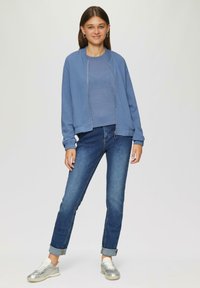 Hellblaues Zip-Up-Sweatshirt mit gerippten Bündchen, kombiniert mit einem passenden Oberteil und dunklen Denim-Jeans. Das Model trägt silberne Sneakers.