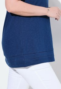 T-Shirt blu navy con vestibilità rilassata, orlo allungato e dettagli di cucitura decorativa sul fondo, abbinato a pantaloni bianchi.