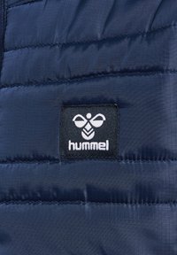 Marineblauwe gewatteerde jas sectie met horizontaal gestikt panelen en een rechthoekig zwart label met een witte bijlogo en "hummel" tekst.