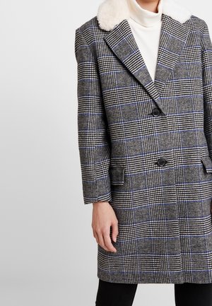 Femme portant un manteau à carreaux gris avec des accents bleus et un col en fausse fourrure blanche, par-dessus un col roulé blanc, debout devant un fond blanc.