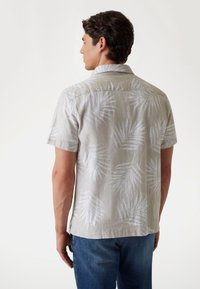 Camisa de manga corta en tela beige clara con patrón de hojas de palmera blancas. Textura suave, diseño con cuello y aberturas laterales en el dobladillo.
