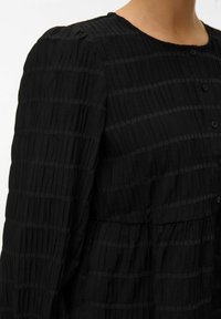 Gros plan d'une personne portant un chemisier noir texturé à manches longues avec des rayures horizontales et verticales et des boutons sur le devant.