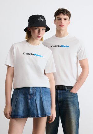 Dwoje modeli w białych koszulkach Calvin Klein; kobieta w dżinsowej spódnicy i czarnym kapeluszu bucket, mężczyzna w niebieskich dżinsach z ręką w kieszeni.