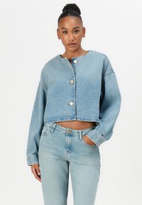 Lichtblauwe denim cropped jas met grote knopen aan de voorkant, verlaagde schouders en een ruwe zoom. Gedragen over bijpassende lichtblauwe jeans.