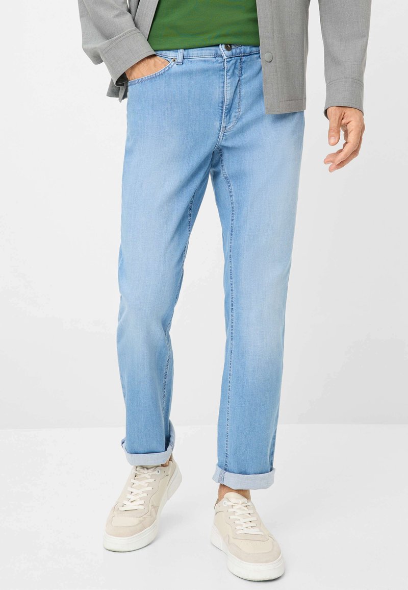 Jeans in denim azzurro chiaro con una vestibilità rilassata, con le maniche arrotolate e tasche frontali, abbinati a sneakers beige e una camicia verde sotto una giacca grigia.
