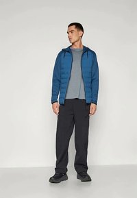 Homem vestindo uma jaqueta com capuz azul, uma camisola cinza, calças cargo pretas e sapatos pretos, em pé contra um fundo claro e liso.