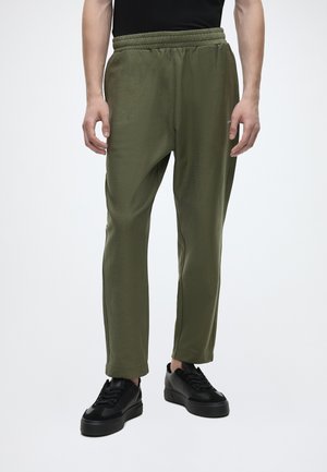Mand iført olivengrønne elastiske bukser med taljebånd og sorte sneakers, stående mod en ensfarvet lys baggrund.