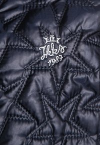 Tessuto trapuntato scuro con cuciture a forma di stella e testo ricamato in bianco "Ilklo 1987" sotto un piccolo simbolo di corona.