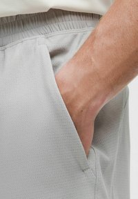 Lichtgrijze shorts van textuurstof, voorzien van zijzakken en een elastische tailleband, waarbij gedeeltelijk een hand in een zak zichtbaar is.
