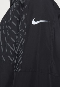 Veste noire avec un motif gris texturé sur l'épaule, arborant un logo Nike blanc bien visible. Tissu léger, texture lisse.