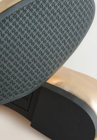 Primer plano de suela de zapato de goma negra con agarre estampado y parte superior metálica dorada con tacón de bloque sobre fondo blanco.