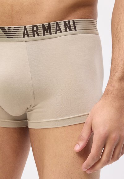 Μπέζ ανδρικά μποξεράκια με το λογότυπο Armani στη μέση, φορεμένα από ένα όρθιο άτομο με το χέρι χαλαρό δίπλα στο μηρό.