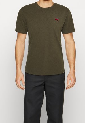 Homme portant un t-shirt à manches courtes vert olive avec un petit camion rouge brodé sur la poitrine, associé à un pantalon noir.