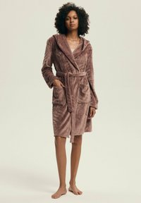 Brązowy pluszowy szlafrok z kapturem, wyposażony w pasek, dwie kieszenie oraz teksturowany wzór. Długość do połowy uda i długie rękawy. Miękki materiał.