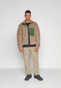 Lichtbruine fleecejas met een groen borstzakje, een grijze rits en een hoge kraag, gecombineerd met beige cargobroek en zwarte schoenen.