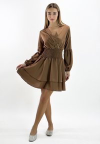 Braunes, langärmeliges Kleid mit V-Ausschnitt, gerafftem Taillenbund und mehrlagigem Rock. Weiche Textur; kombiniert mit silbernen Ballerinas.