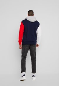 Sudadera con bloques de color en secciones de azul marino, rojo, blanco y gris, con capucha gris, combinada con jeans oscuros y zapatillas blancas.