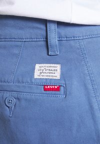 Gros plan sur la poche d'un pantalon Levi's bleu clair avec bouton, détails cousus, étiquettes Levi's blanches et rouges, et une partie d'une chemise blanche au-dessus.