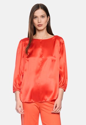 Woman with long brown hair wearing a bright red satin blouse with three-quarter sleeves and matching red pants, standing against white background.