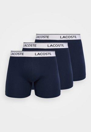 Drie marineblauwe herenboxershort met witte elastische taillebanden met het merk "LACOSTE" opgestapeld op een witte achtergrond.