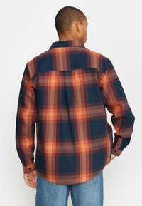 Camisa de manga larga con un patrón de cuadros en azul marino y naranja, confeccionada en tela tejida con dobladillo curvado y puños abotonados.